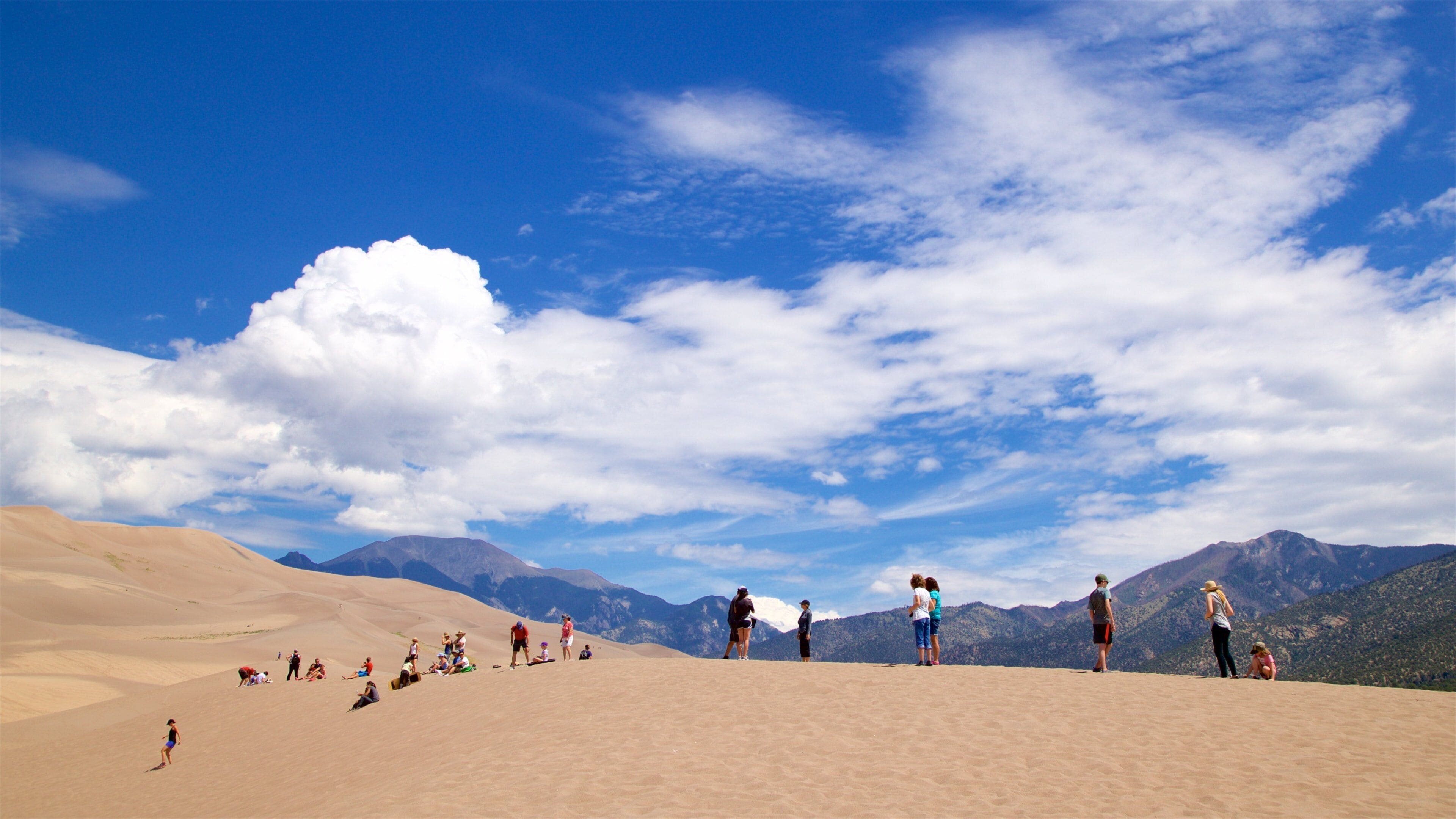 A desert landscape with sand dunes and mountains in the background under a partly cloudy sky.