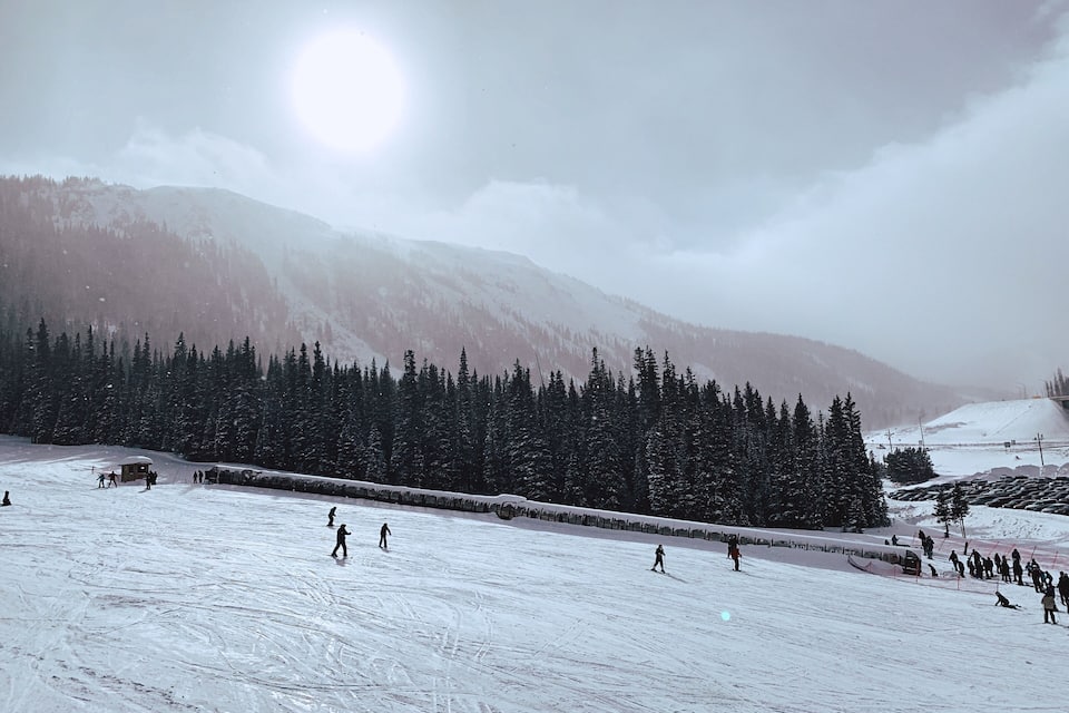 A snowy ski slope with skiers, a mountain backdrop, and a bright sun.