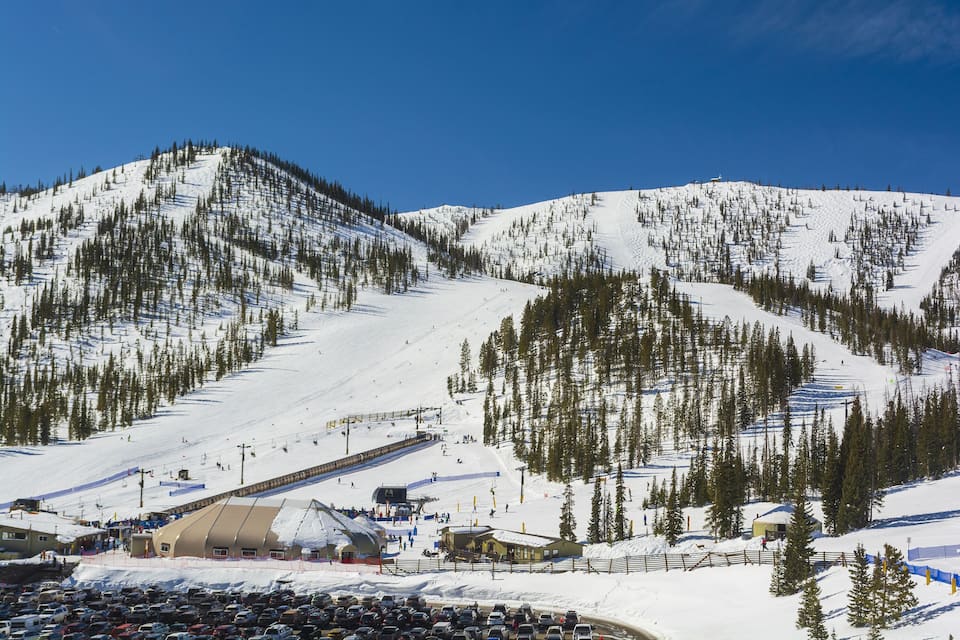 A snow-covered ski slope with a ski lift, a parking lot, and a lodge.