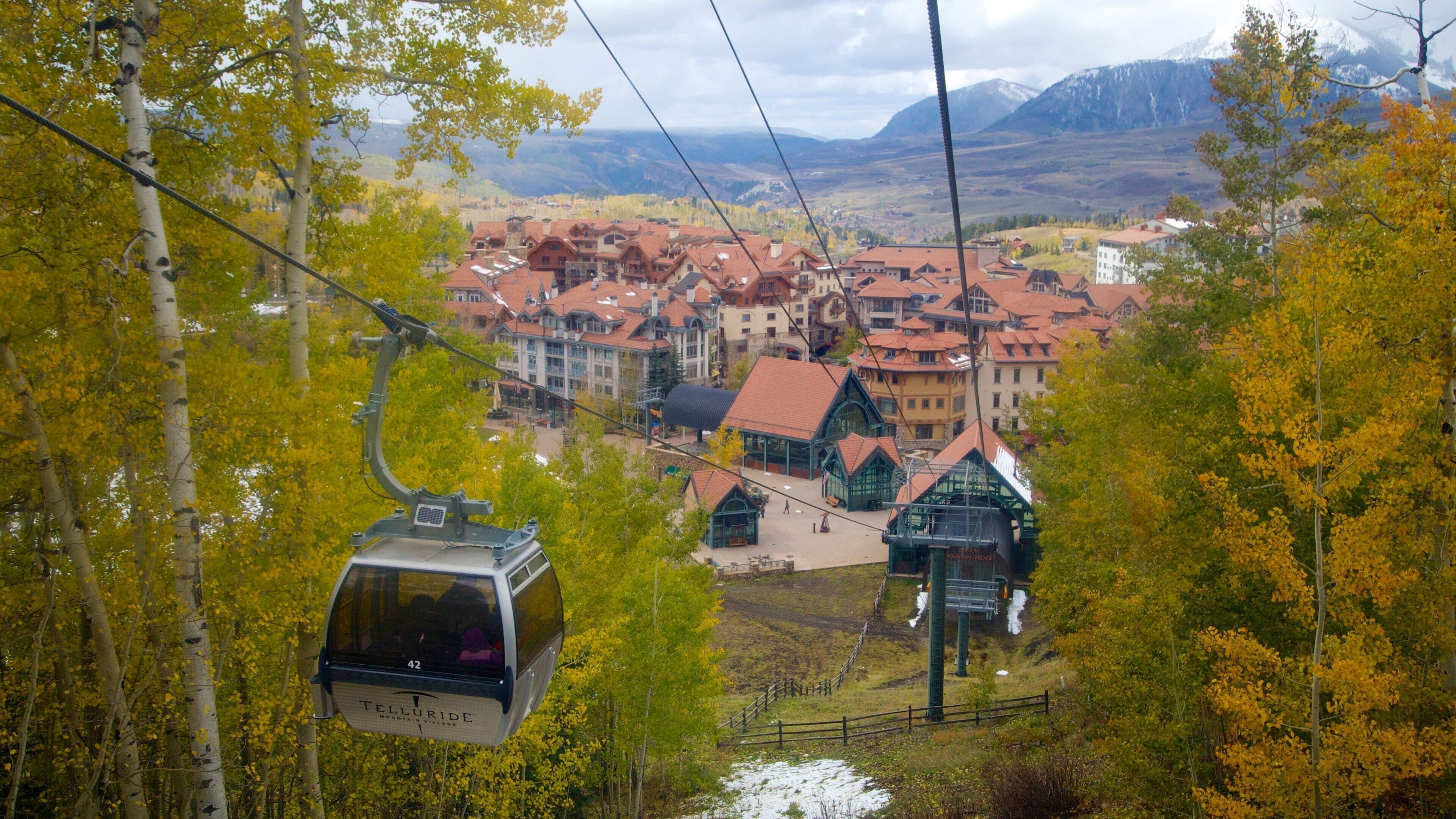 A cable car in a mountainous area with autumn-colored trees and a village with red-roofed buildings.