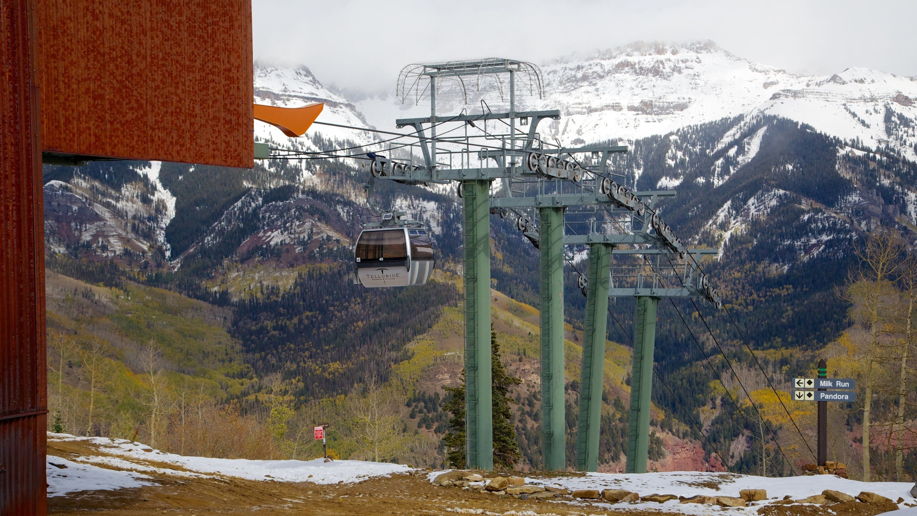 A chairlift in a mountainous area with snow-covered peaks and a sign indicating directions to 'Milk Run' and 'Pandora'.