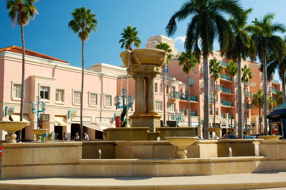 A fountain with a large urn in the center, surrounded by palm trees and a pink building with balconies.