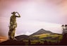 Eine Statue einer Figur mit Schwert, die über eine gebirgige Landschaft mit grünen Feldern blickt.