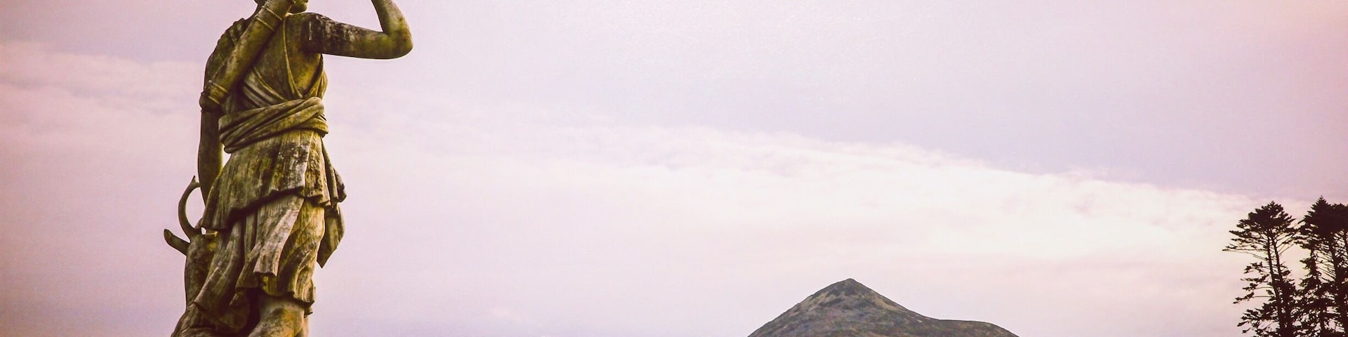 Estátua de uma figura com espada, com vista para uma paisagem montanhosa com campos verdes.