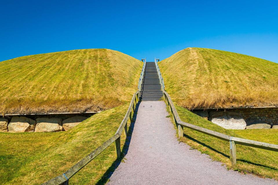 Ein Weg führt zu zwei grasbewachsenen Erhebungen mit einer Holztreppe und Geländern.
