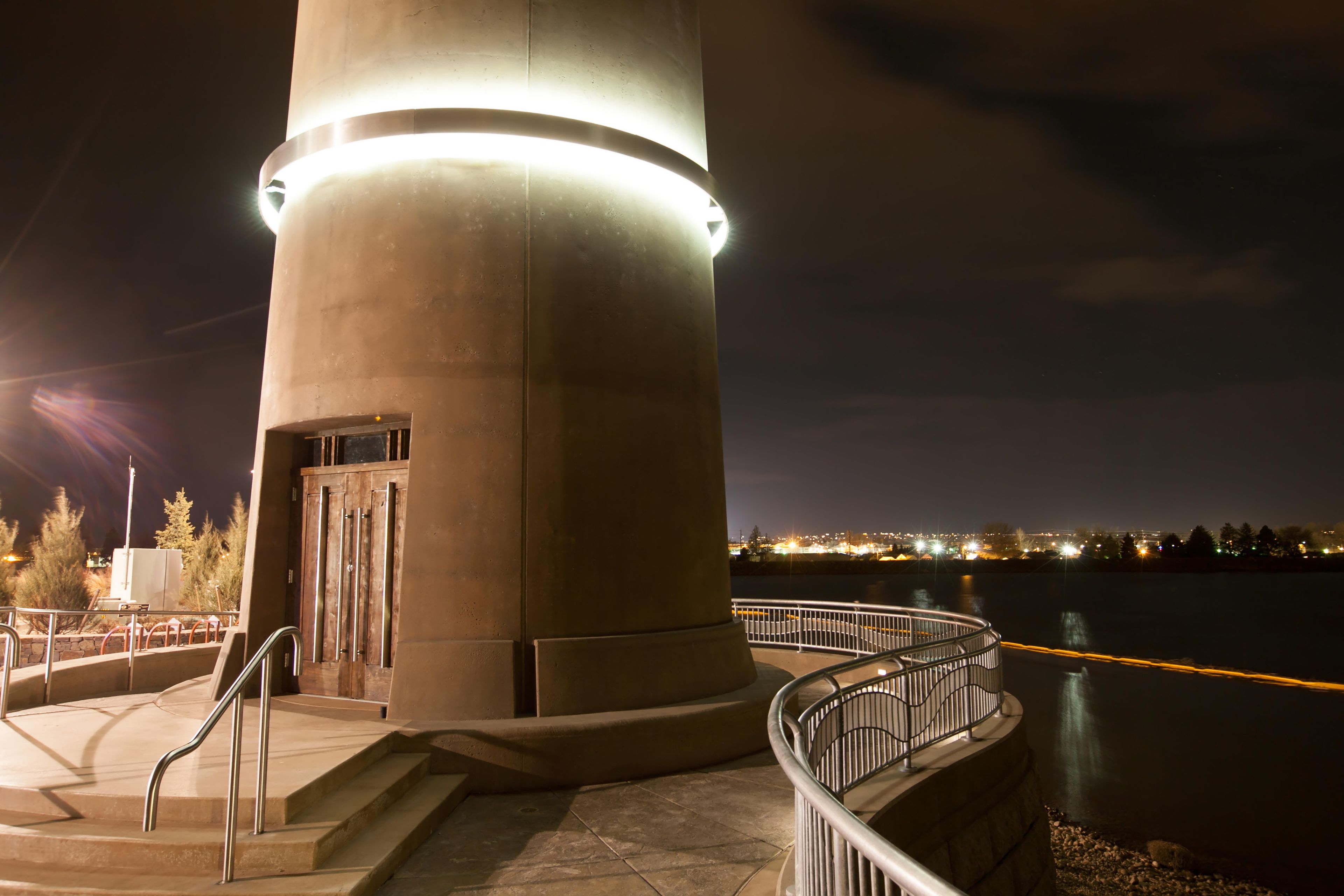 A large concrete structure with a staircase and railing, overlooking a body of water with city lights in the distance.