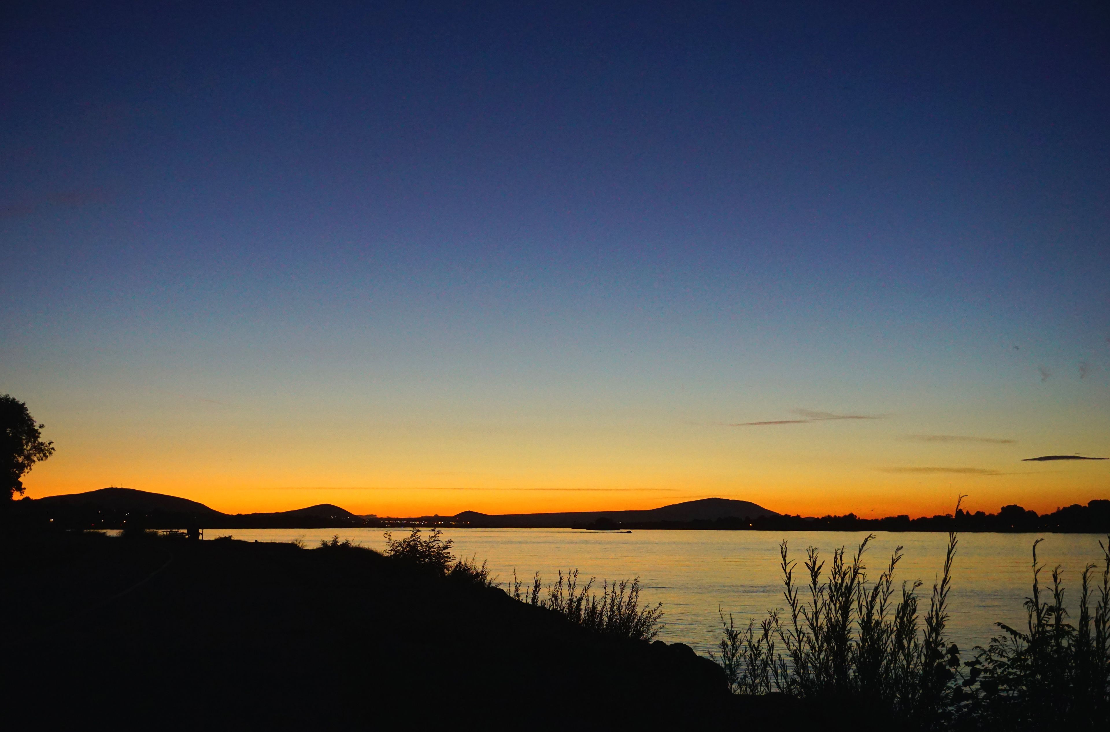 A sunset over a calm lake with silhouettes of hills and sparse vegetation.