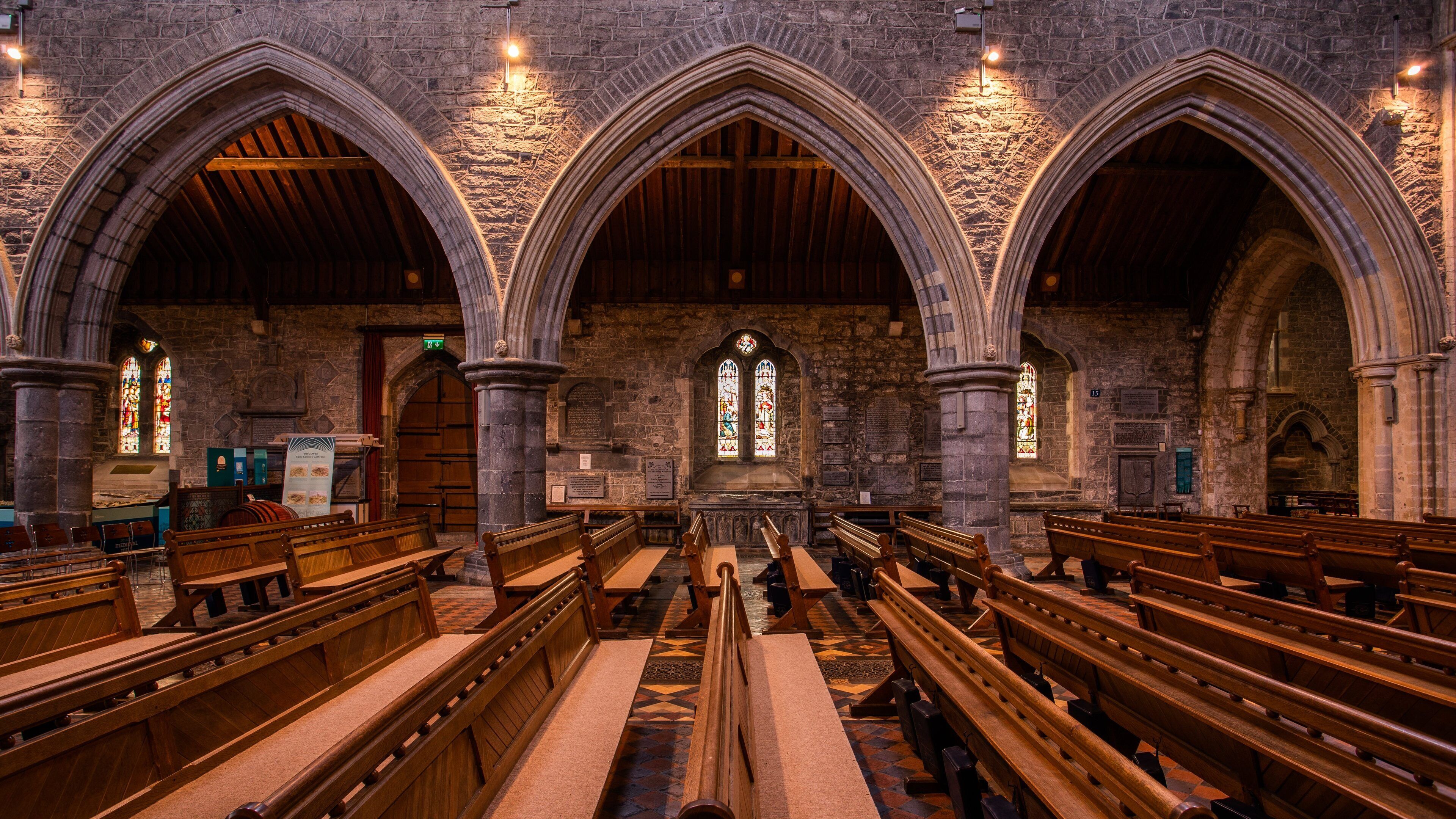 A church interior with wooden pews, stone walls, and stained glass windows.