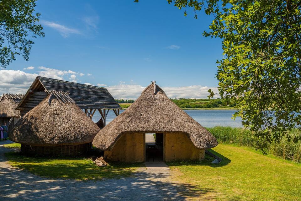 Thatched-roof huts by a lake, a wooden structure, and a clear blue sky.