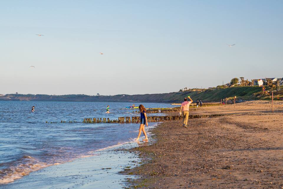 A beach with people flying kites, a rocky shoreline, and houses in the background.