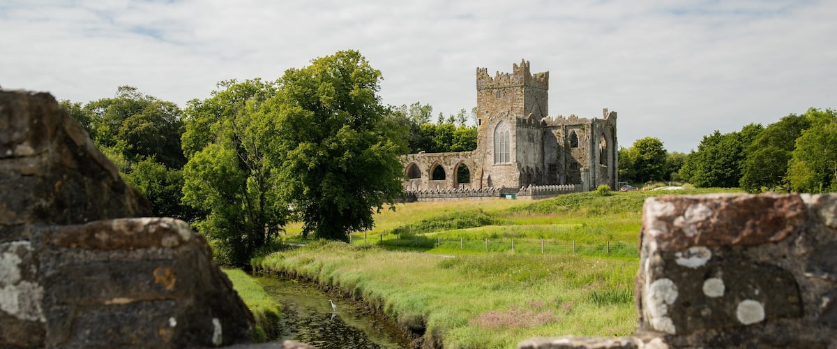 A historic stone castle with arched windows and a tower, surrounded by lush greenery and a narrow stream.