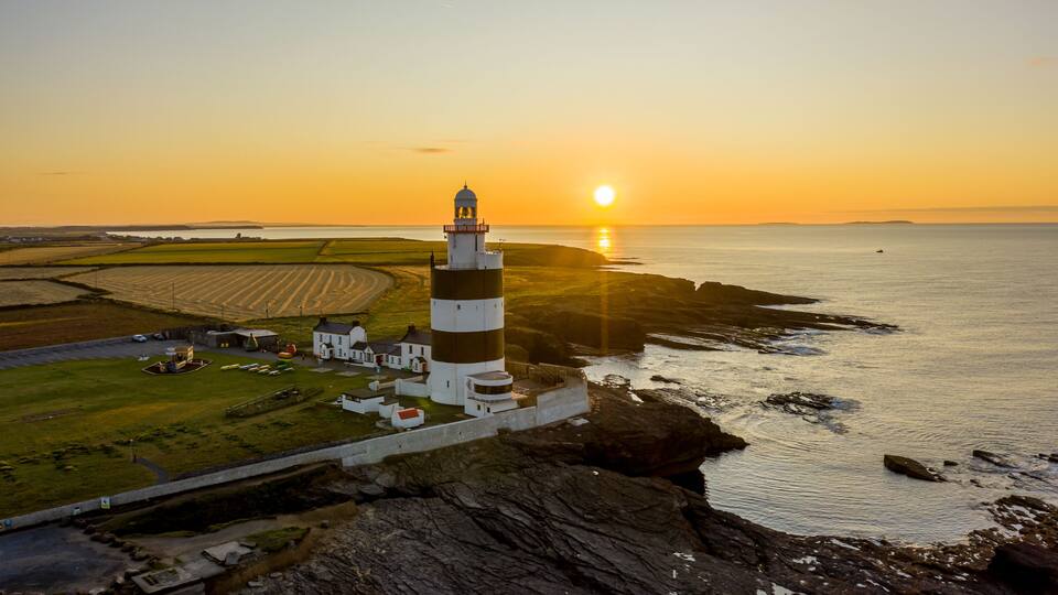 A lighthouse on a rocky coastline with fields in the background and the sun setting over the horizon.