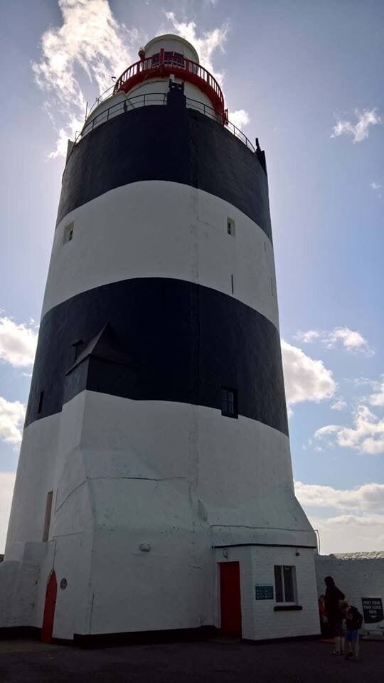 A tall, cylindrical lighthouse with alternating black and white stripes, a red railing at the top, and a small window at the base.