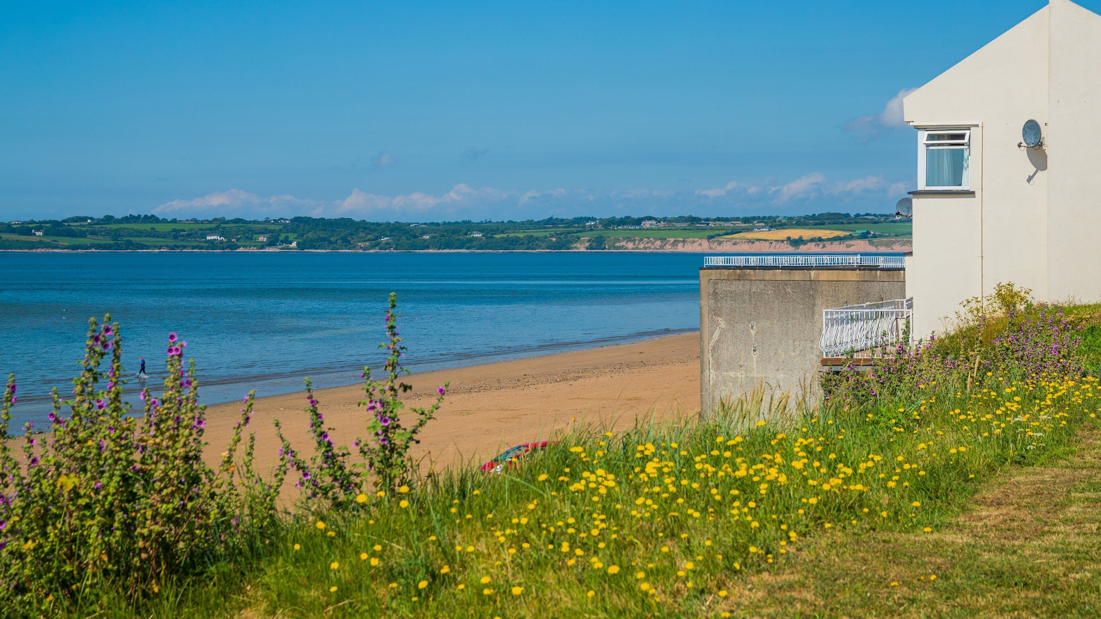 A coastal landscape with a sandy beach, a white building with satellite dishes, and a clear blue sea.