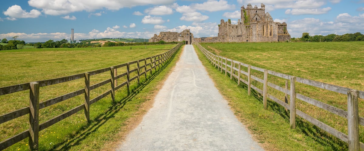 A historic castle with a paved pathway leading to it, surrounded by green fields and a clear sky.