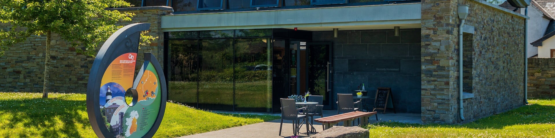 A modern building with a glass facade, outdoor seating, and a large informational sign.