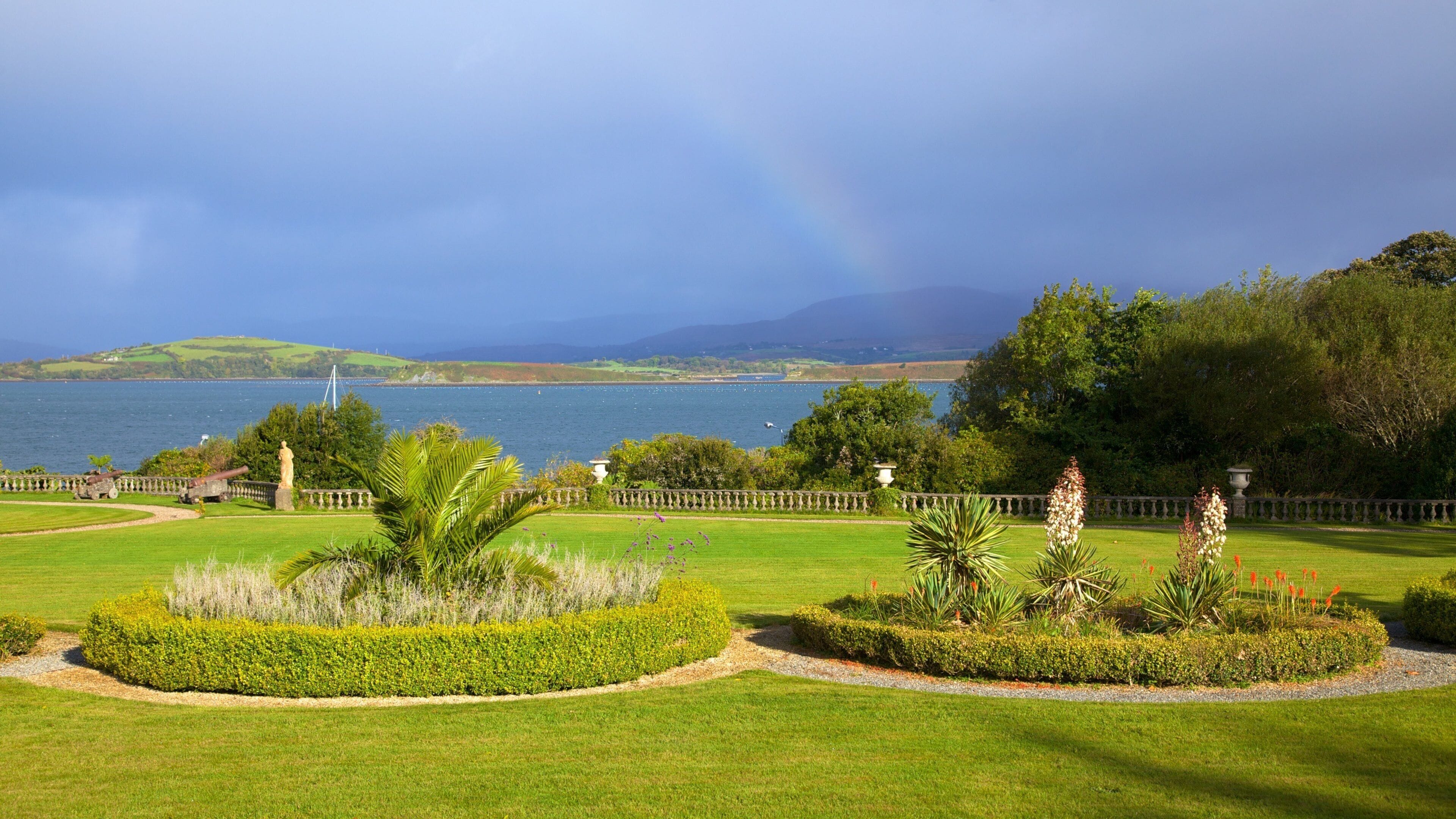 A landscaped garden with manicured lawns, a statue, and a view of a lake with mountains in the background.