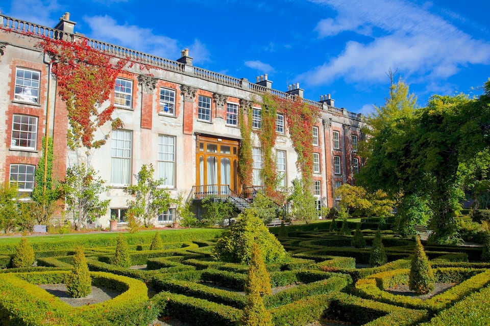 A historic building with red brick and white stone, surrounded by a formal garden with geometric hedges and mature trees.