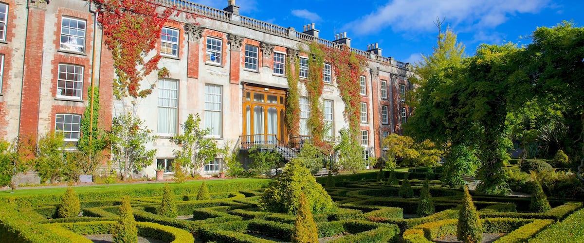 A historic building with red brick and white stone, surrounded by a formal garden with geometric hedges and mature trees.