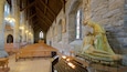 An interior of a church with wooden pews, stone walls, and a statue of a woman kneeling.