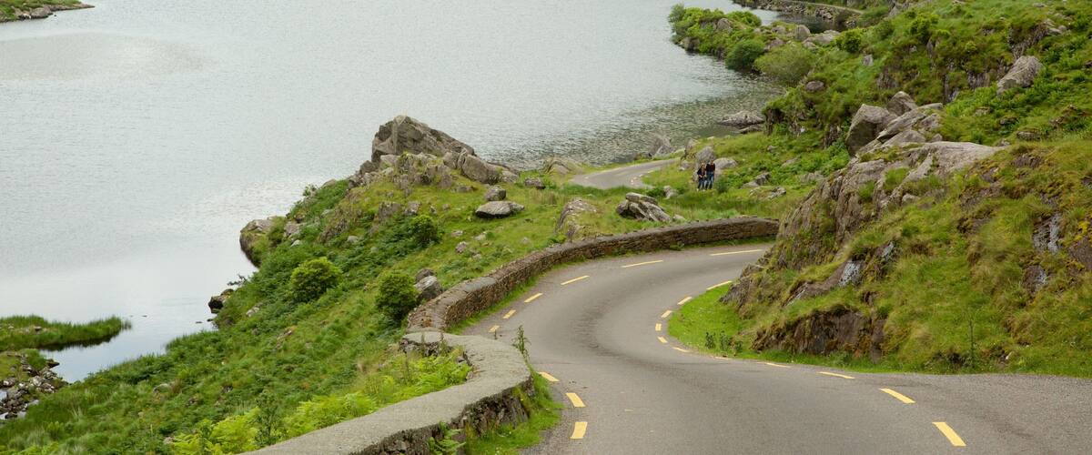 A winding road alongside a lake with rocky terrain and greenery.