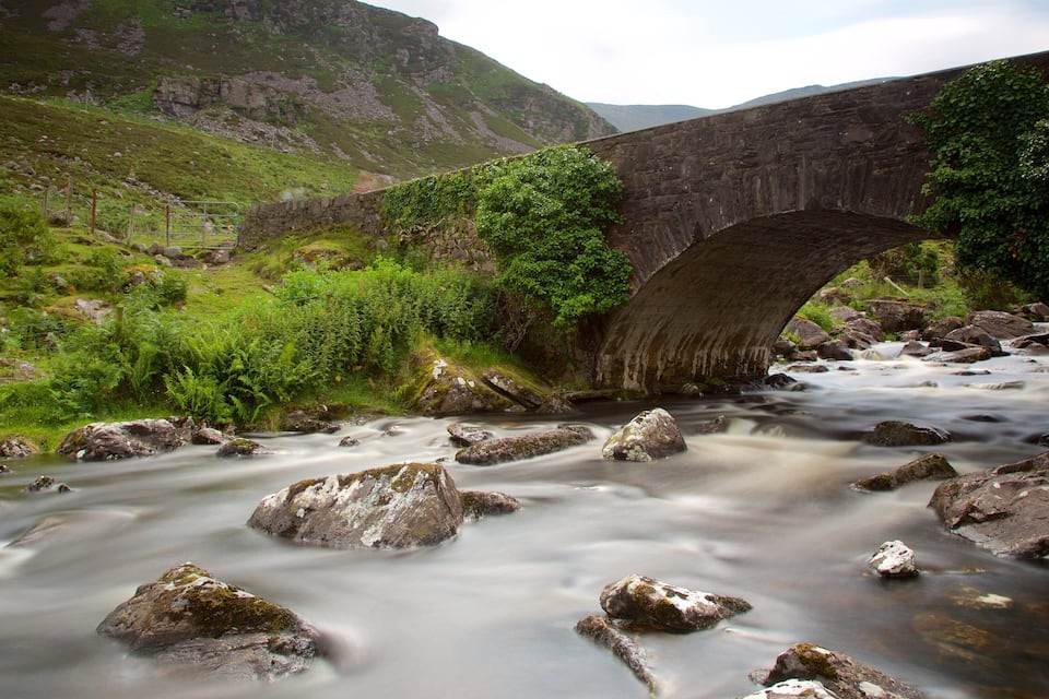 A stone bridge over a flowing river with surrounding greenery and hills.