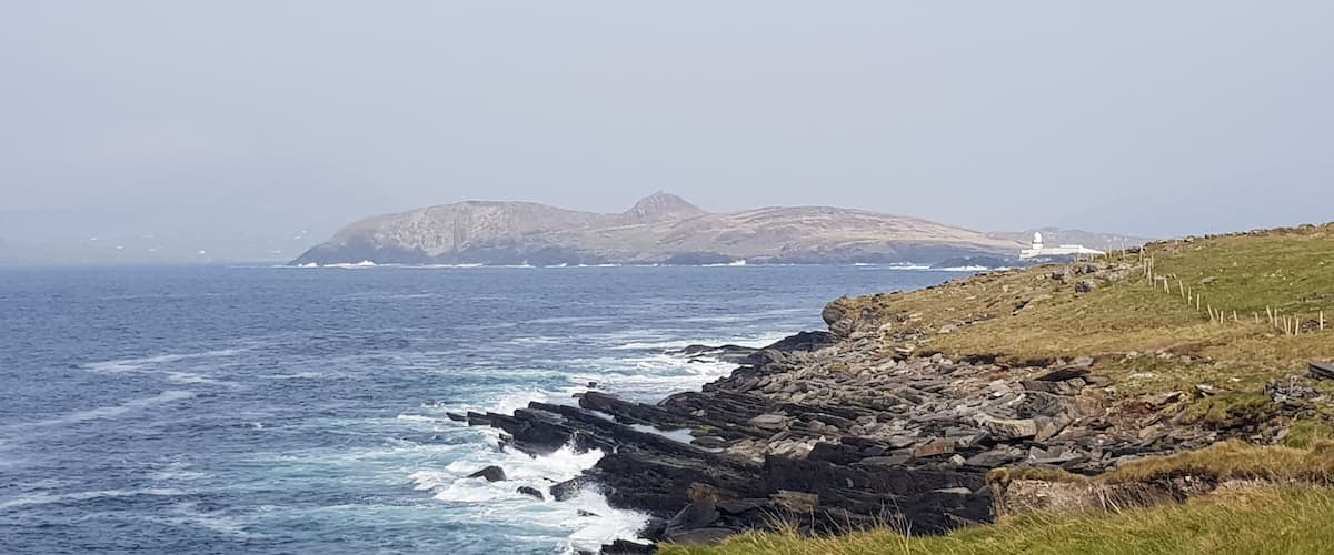 A rocky coastline with waves crashing against the shore, a lighthouse in the distance, and a mountain range in the background.