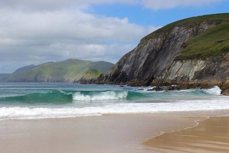 Une plage côtière avec des vagues, un rivage sablonneux et une falaise rocheuse parsemée de végétation.