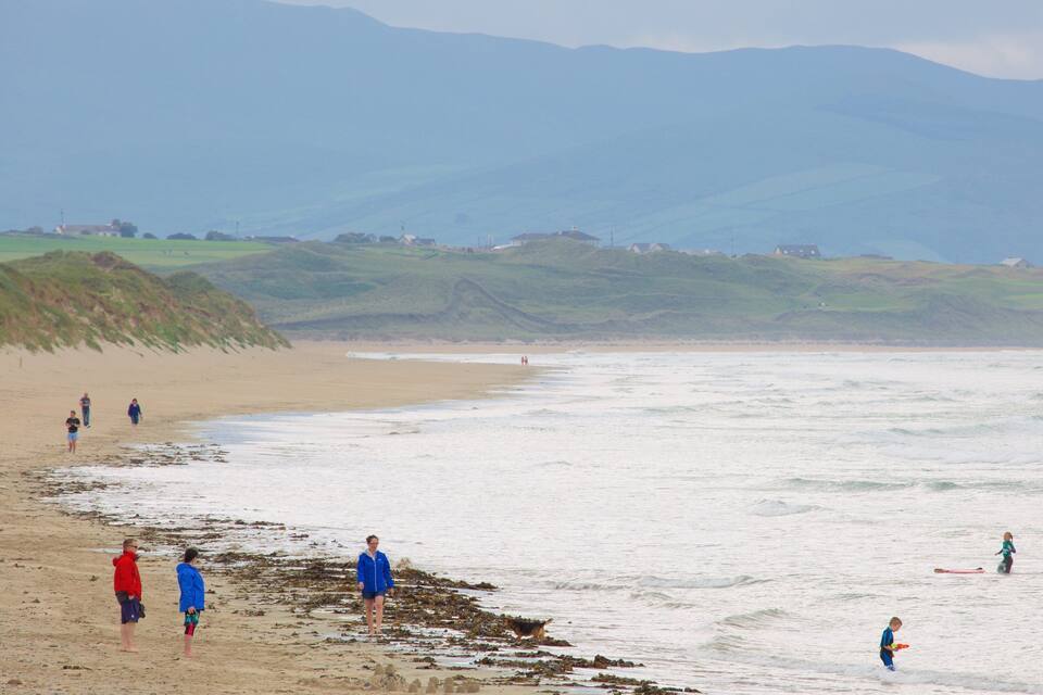 Una playa con gente caminando y jugando en el agua, rodeada de colinas y casas al fondo.