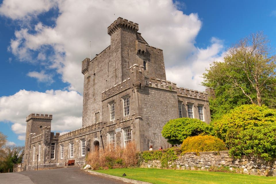 A large stone castle with a prominent tower, surrounded by lush greenery and a clear blue sky.