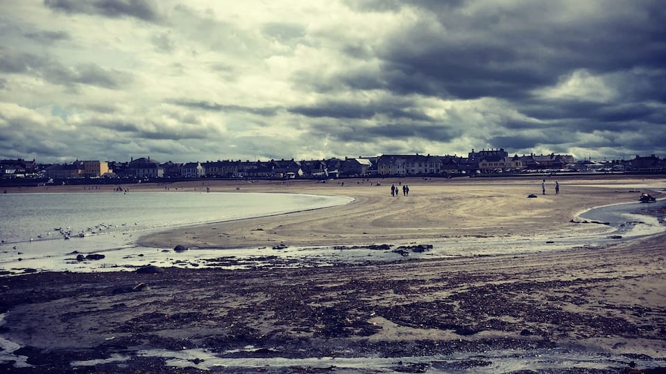 A beach with a cloudy sky, a sandy shore, and buildings in the background.