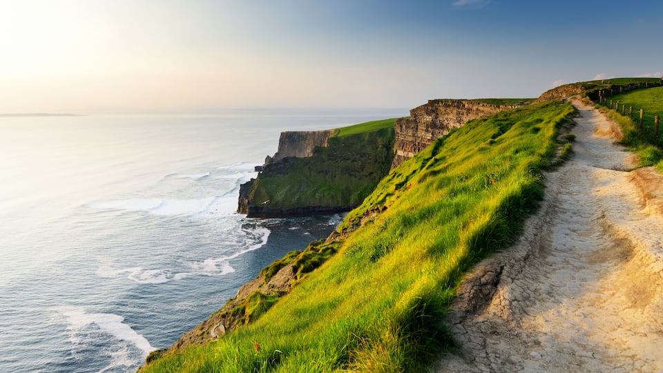 A coastal path with cliffs, ocean waves, and a grassy hillside.