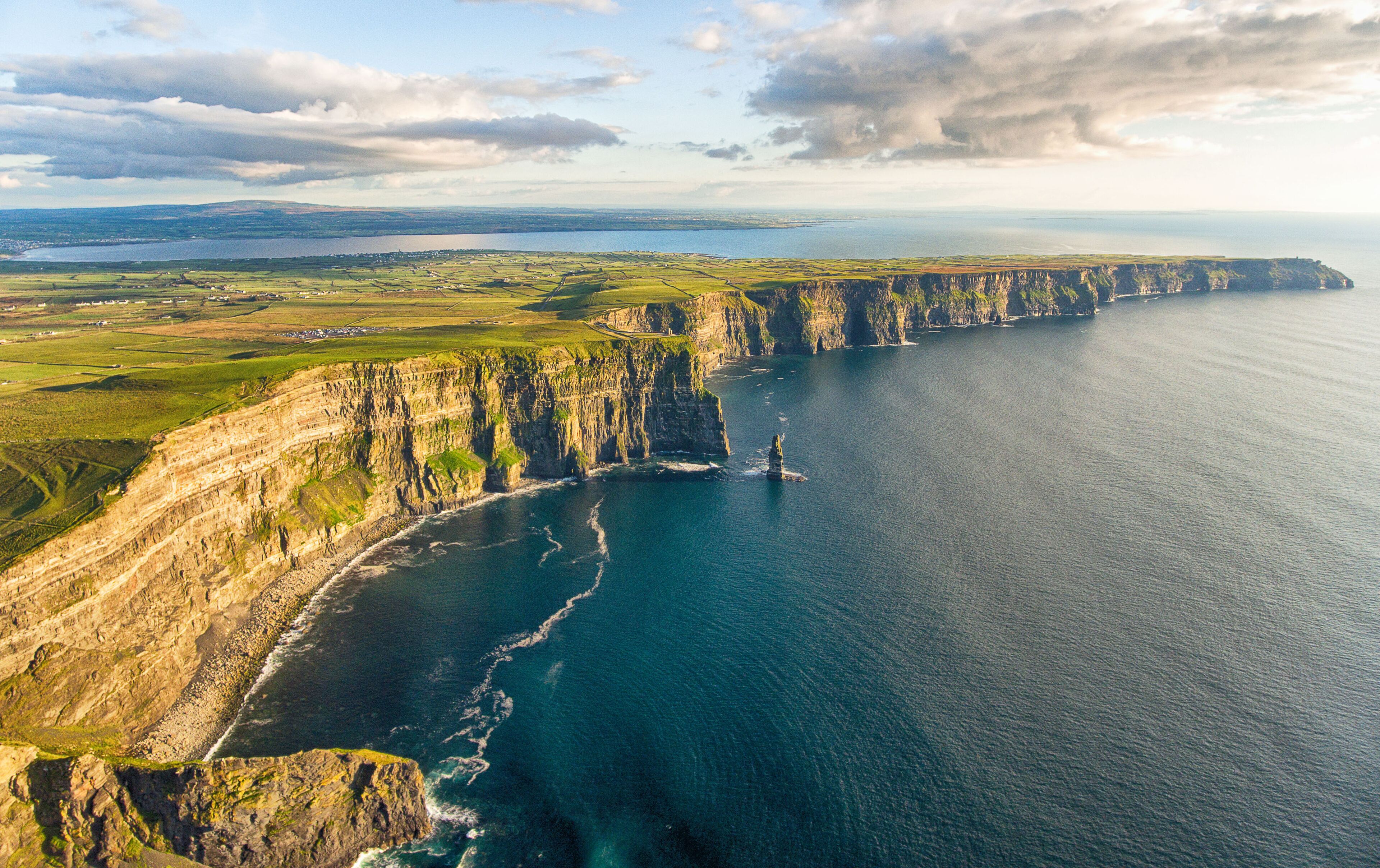 A coastal cliff with a narrow sea passage, a distant shoreline, and a clear sky.
