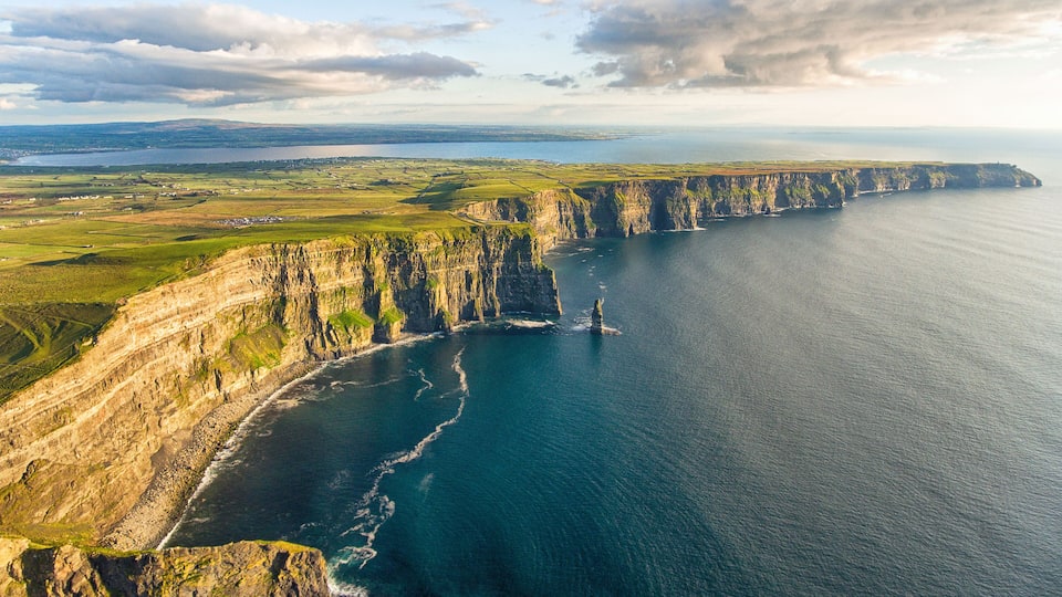 A coastal cliff with a narrow sea passage, a distant shoreline, and a clear sky.