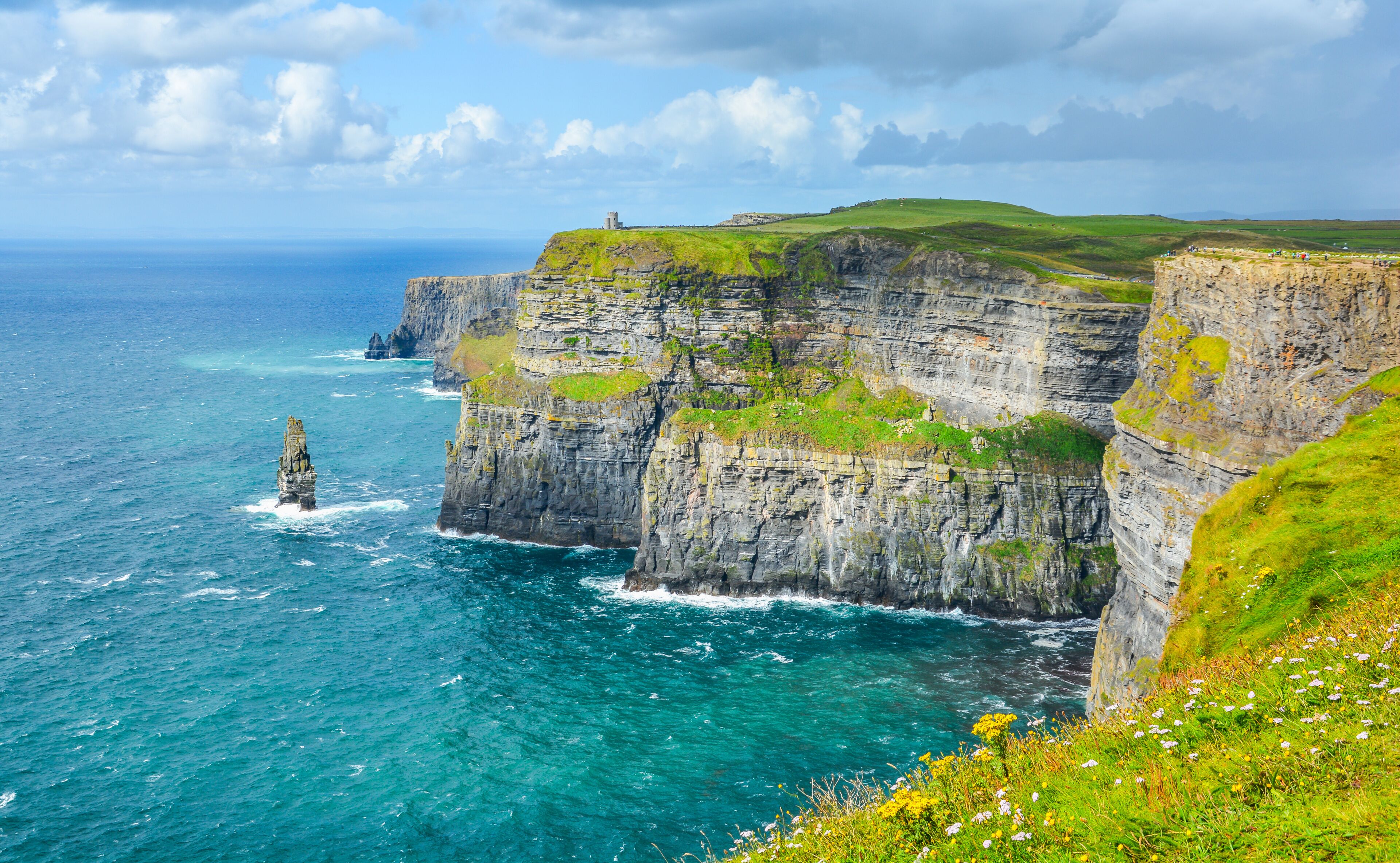 A coastal cliff with a lighthouse, ocean waves, and grassy terrain.