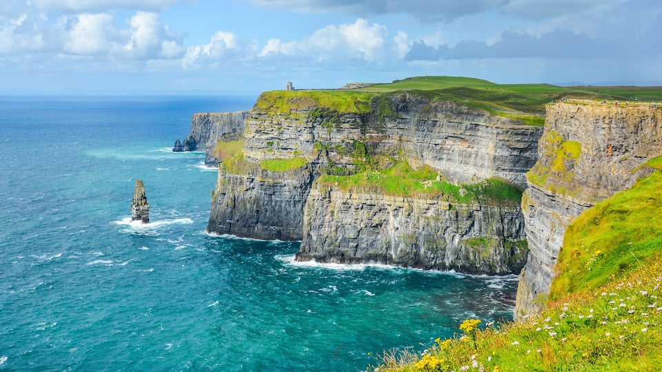 A coastal cliff with a lighthouse, ocean waves, and grassy terrain.