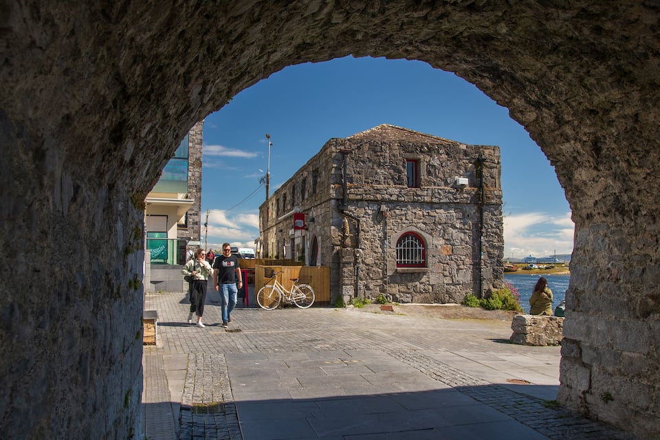 Una galeria d'arcs emmarca un edifici històric de pedra amb una finestra vermella i una placa.