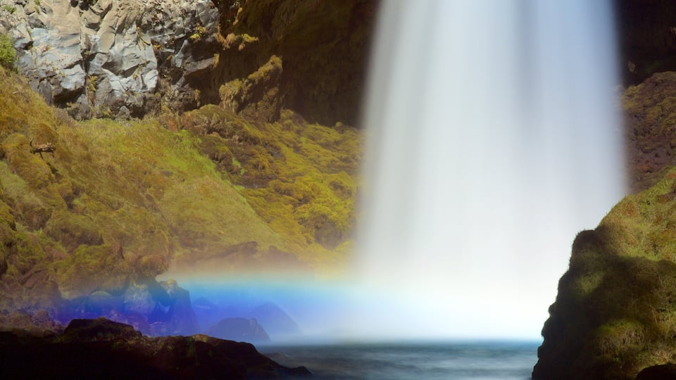 A waterfall with a rainbow at its base, surrounded by rocky terrain and greenery.