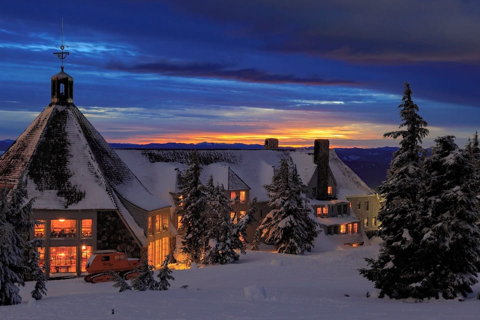 Een huis bedekt met sneeuw, met een opvallende schoorsteen, en een besneeuwd landschap in de zonsondergang.