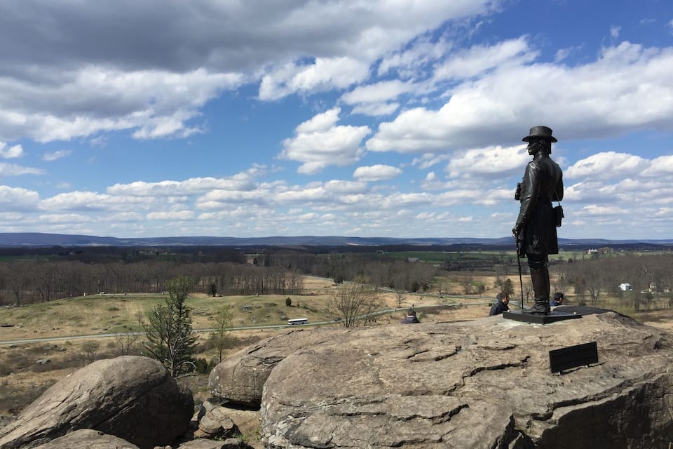 A statue on a rocky outcrop overlooking a valley with distant hills and a partly cloudy sky.