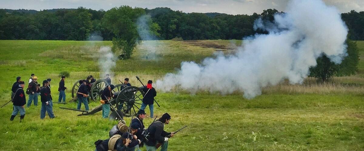 A historical reenactment with soldiers in period uniforms, a cannon, and smoke in a grassy field under a cloudy sky.