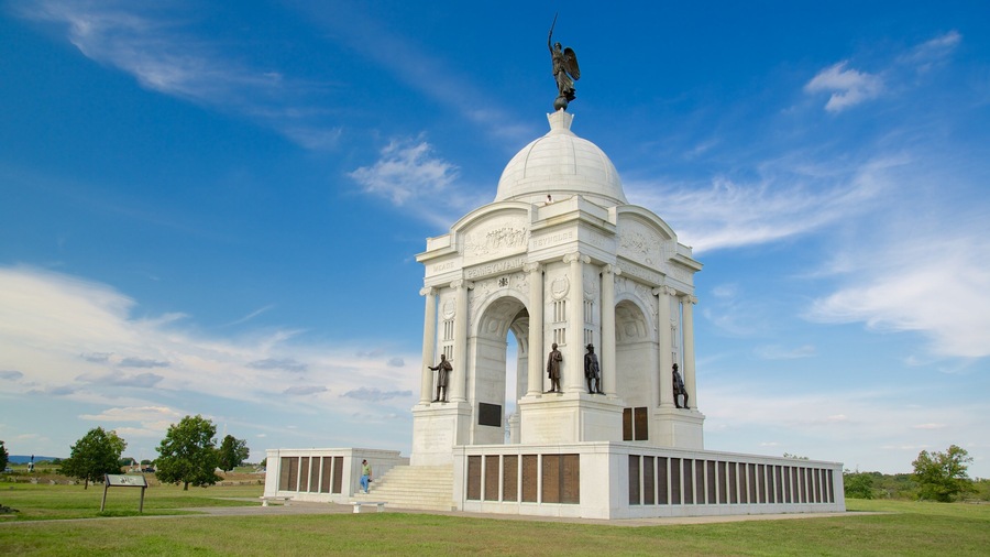 Un monument blanc orné d’inscriptions et de statues, entouré d’un champ verdoyant et surplombé d’un ciel bleu clair.