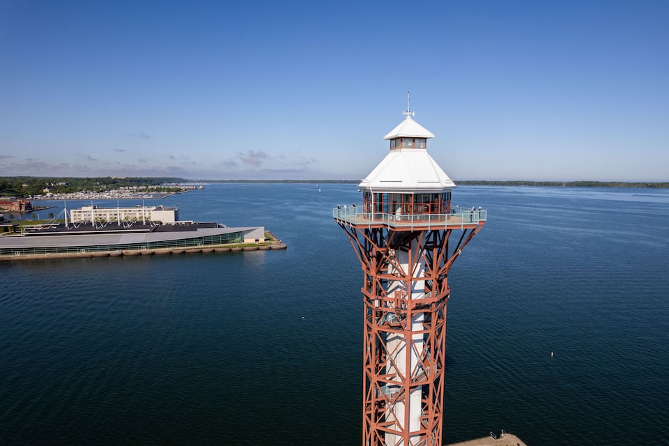 A coastal tower with a white dome and red metal structure, overlooking a calm sea and a marina with boats.