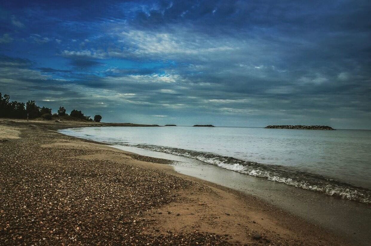 A pebbly beach with calm waters and distant islands under a cloudy sky.