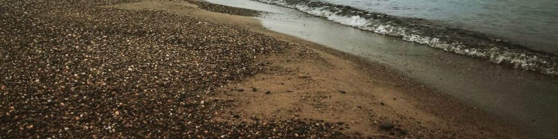 A pebbly beach with calm waters and distant islands under a cloudy sky.