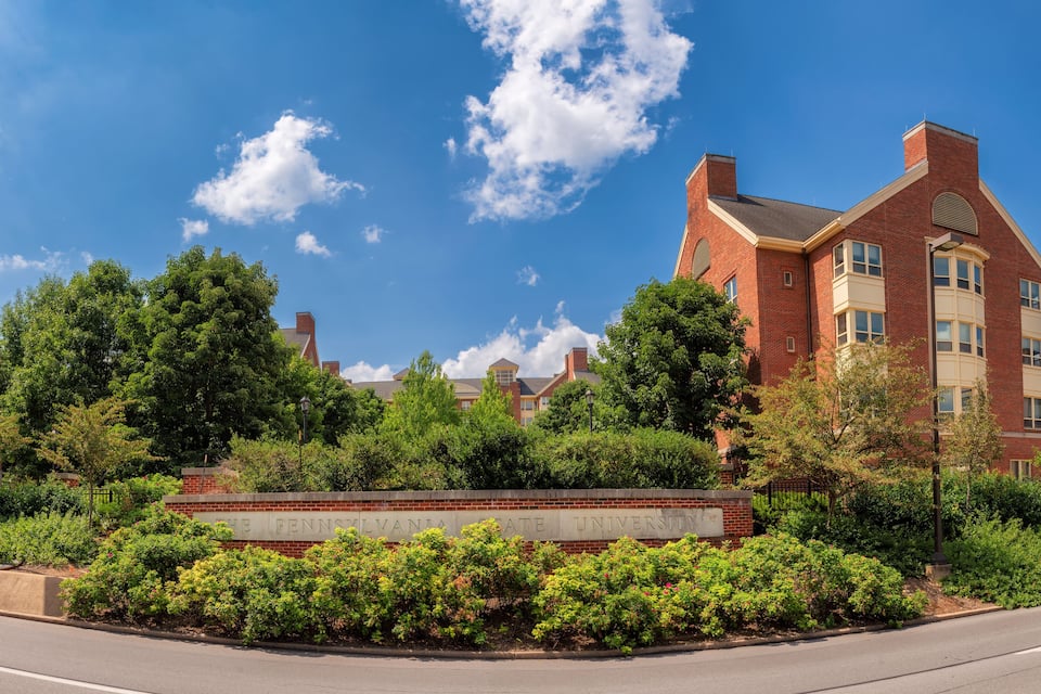 Um edifício de tijolos com uma placa que diz "The Pennsylvania State University", cercado por vegetação e um céu azul claro.