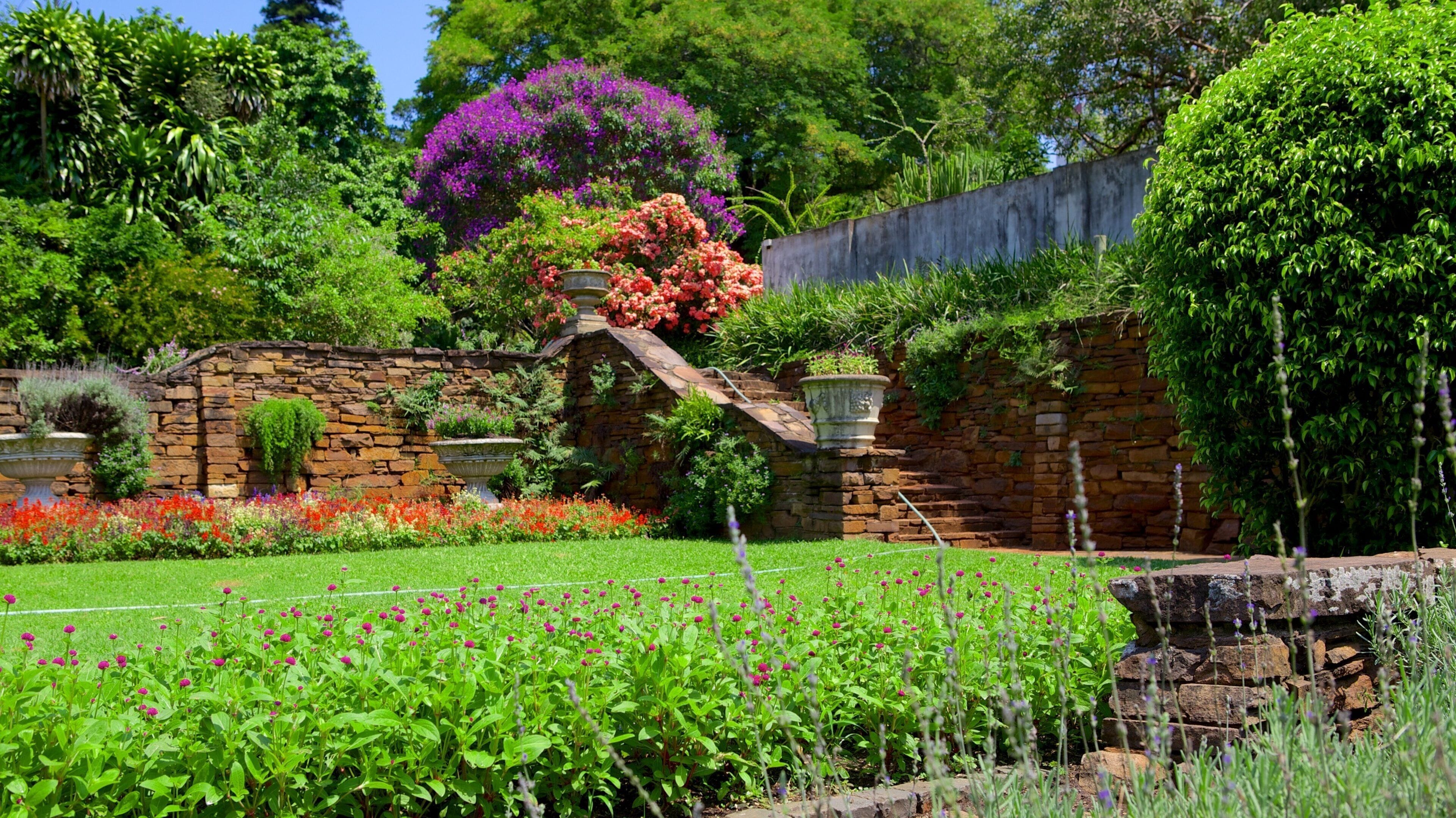 A lush garden with a stone wall, flowering plants, and a variety of trees.