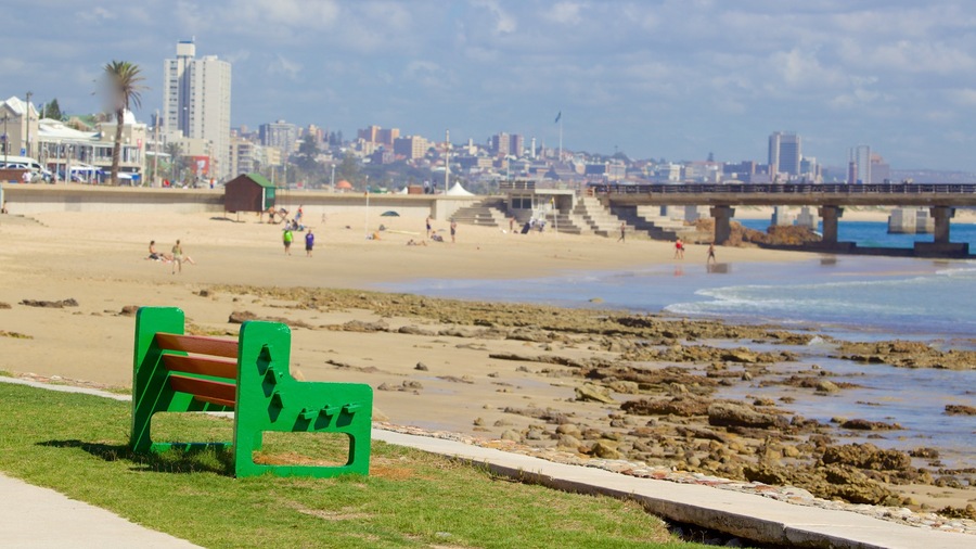 A beach with a green bench, rocky shoreline, and a cityscape in the background.