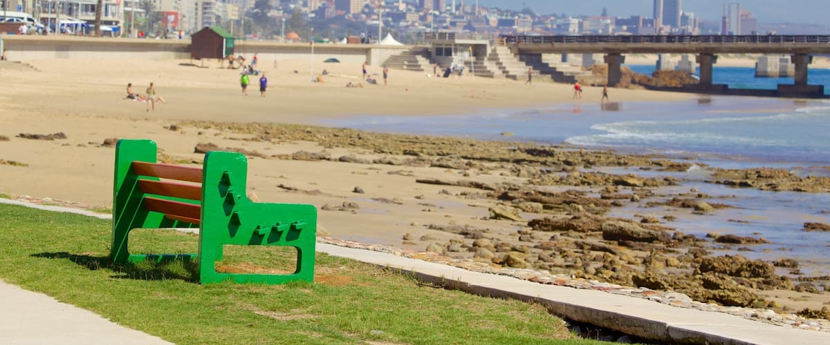 A beach with a green bench, rocky shoreline, and a cityscape in the background.