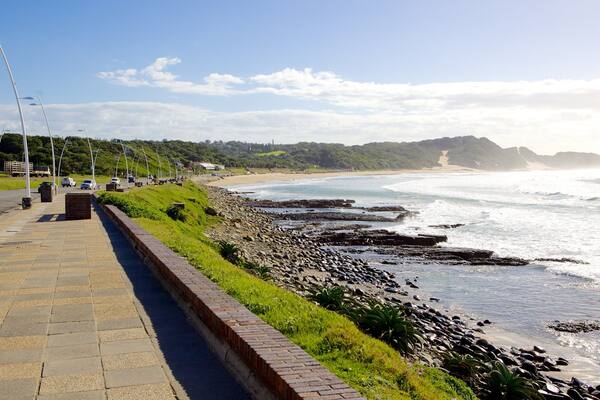 A coastal walkway with a rocky shoreline, a clear sky, and distant hills.